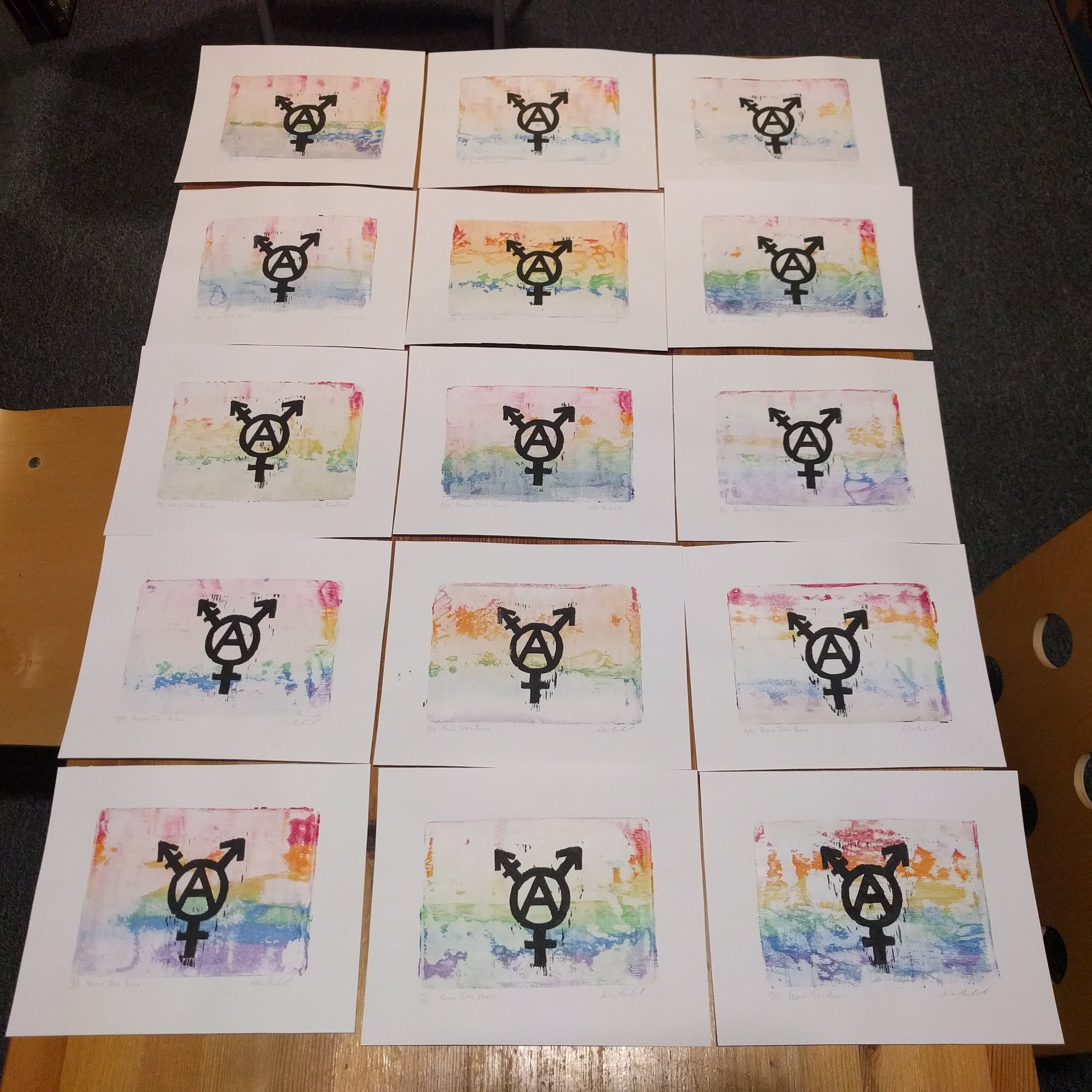 15 linoprints of a trans anarchy symbols in black on a background of a ghost gelprints of a horizontal rainbow gradient, laid out so that the range of rainbow effects are visible.