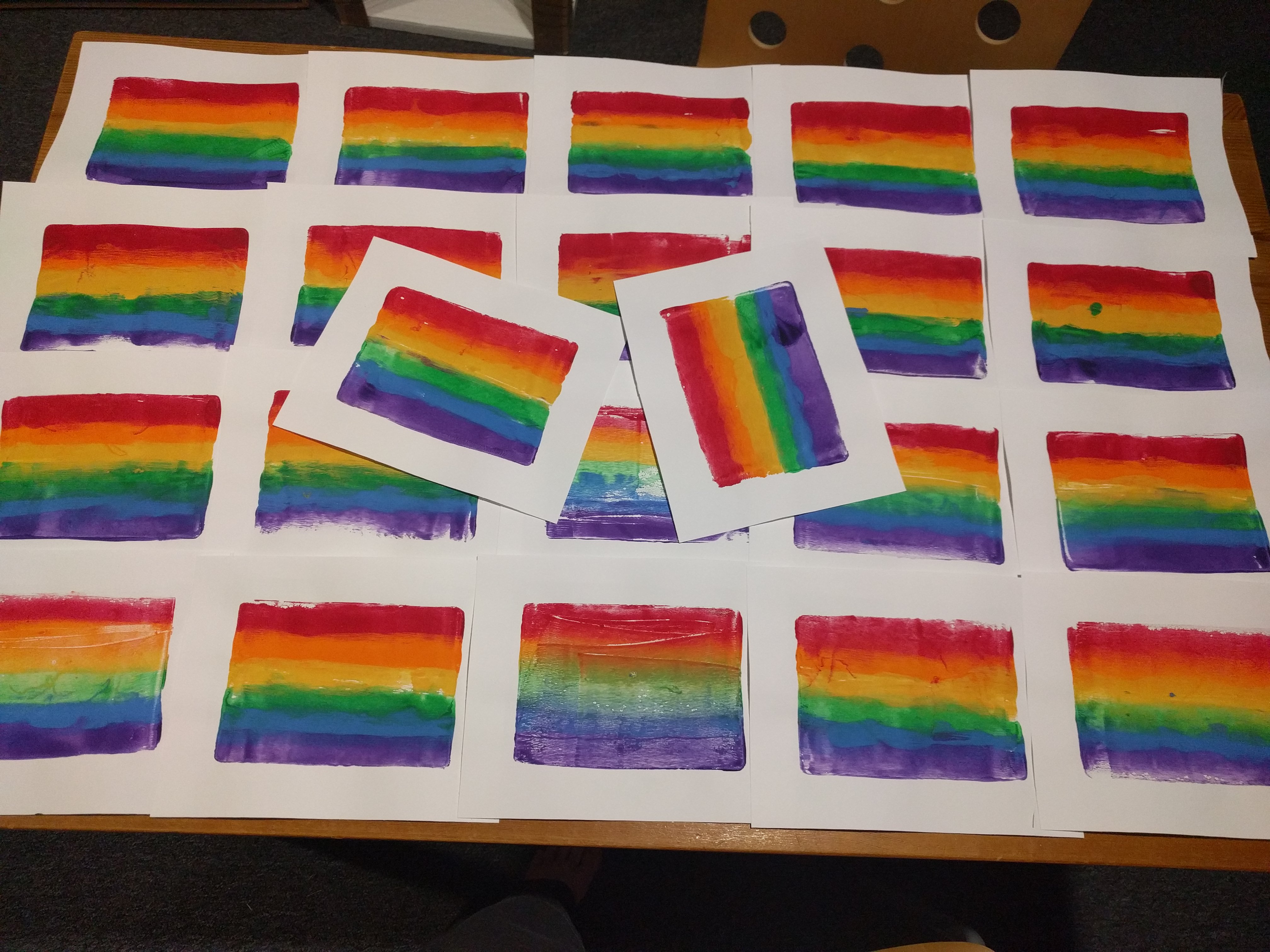 A tablefull of gelprints on white paper, each with a rainbow gradient from top to bottom. There is an orderly four by five grid, with an additional two tossed into the center haphazardly 