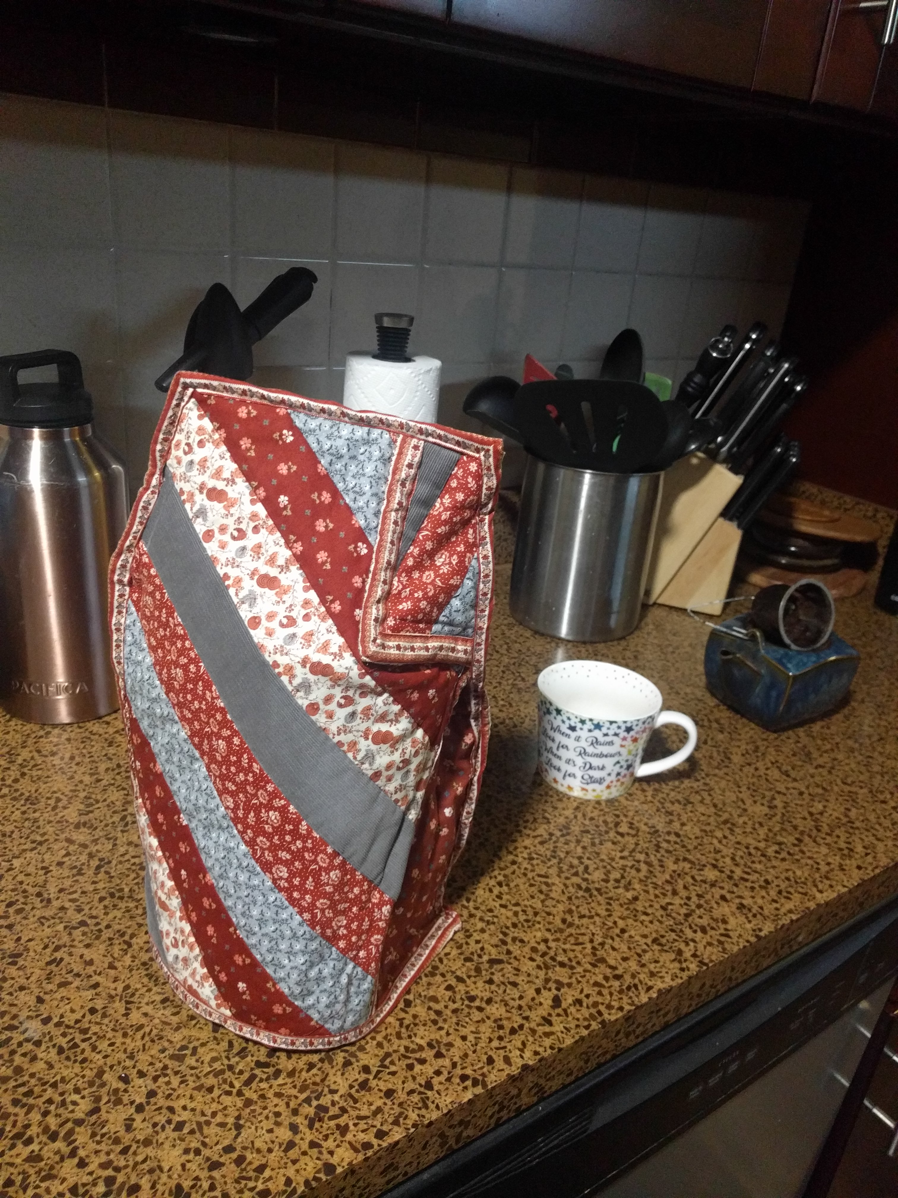 The scene of the french press from the beginning of this page, now with a striped, quilted, burgundy and blue cozy on top of it