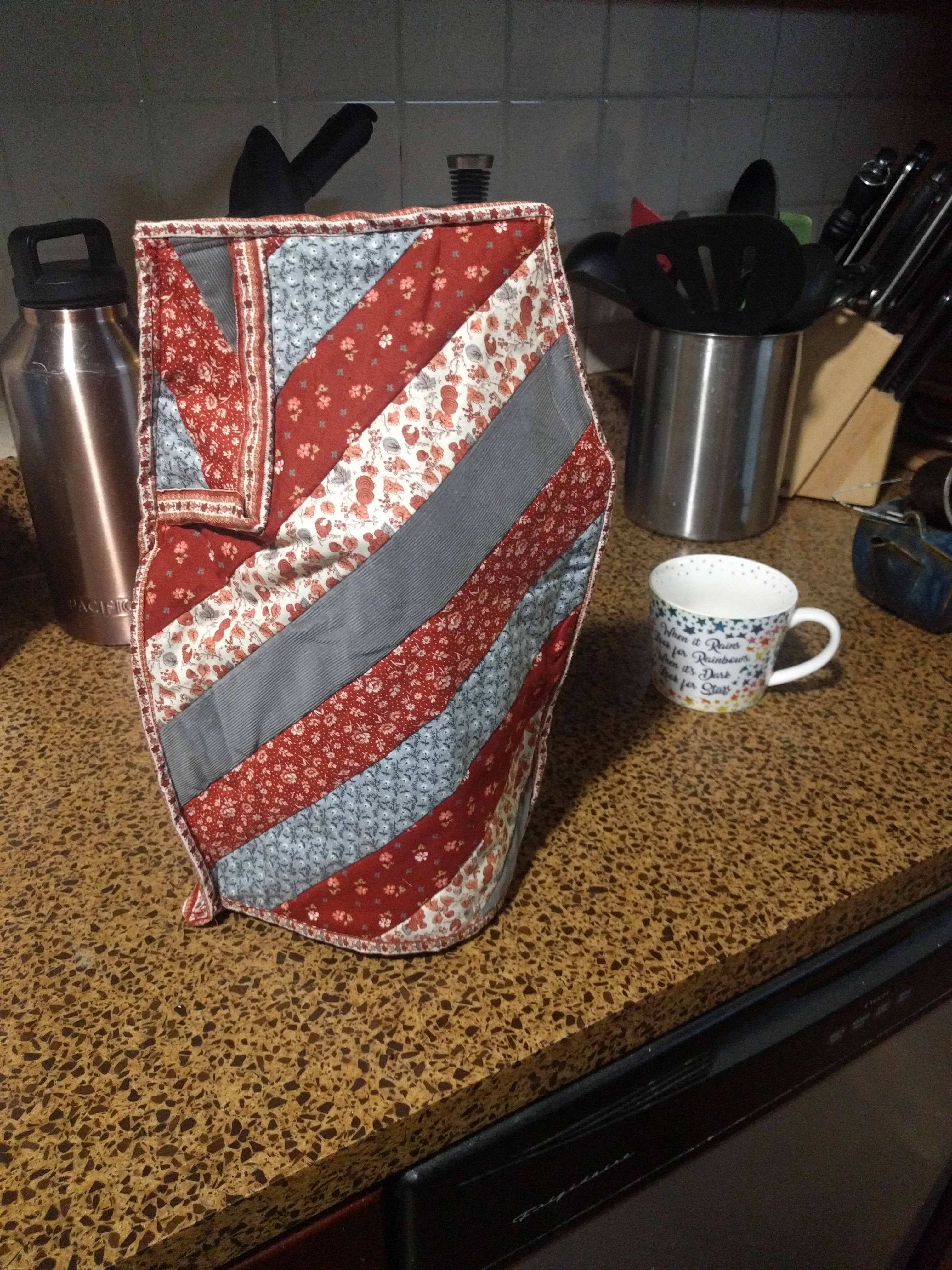 The scene of the french press from the beginning of this page, now with a striped, quilted, burgundy and blue cozy on top of it, turned 180 degrees, which highlights the piecing differently