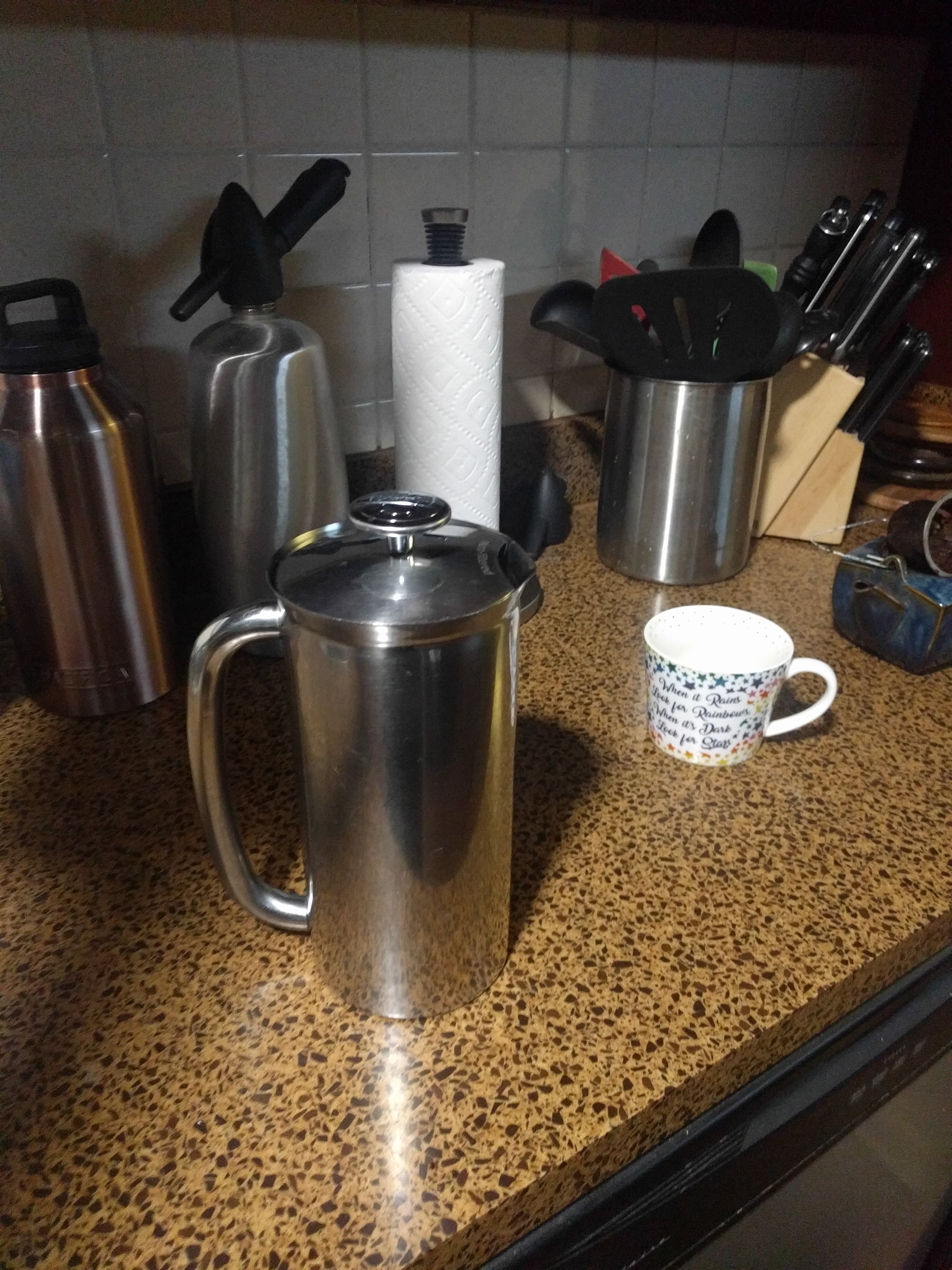 A stainless steel large french press sitting on a countertop. Behind the french press is a variety of kitchen implements, a knife block, a selzer bottle and paper towels on a holder amongst them.