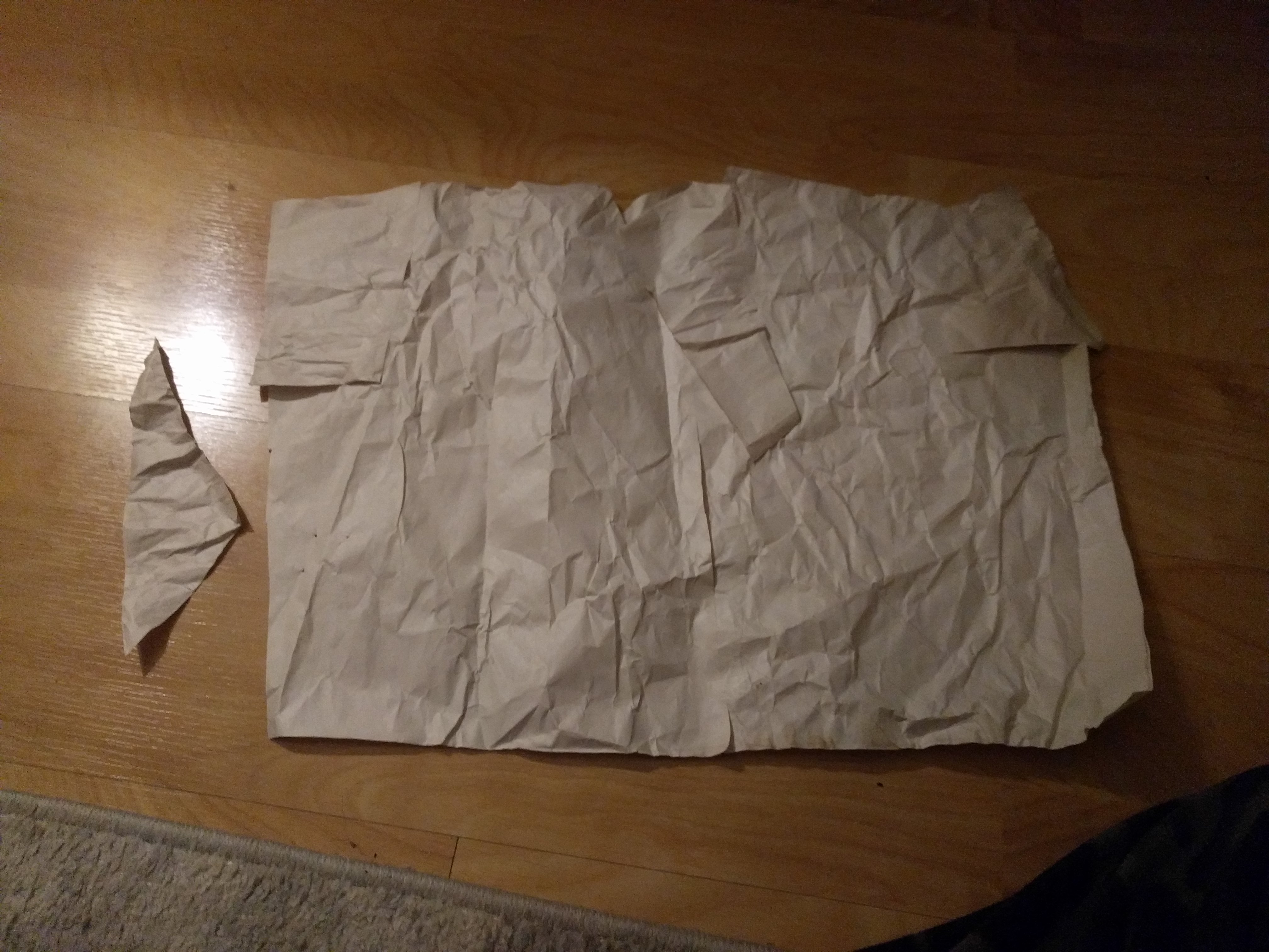 An approximately rectangluar piece of offwhite, wrinkled paper that at closer inspection is actually several smaller pieces stuck together with tape. One small triangle of paper is off to the side, having been not necessary to create the rectangle. Both pieces of paper are laying on a wooden floor